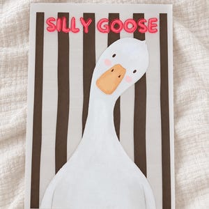 Puede incluir: Una tarjeta rectangular que muestra un ganso blanco con la cabeza inclinada, pico naranja y mejillas rosadas. El fondo consiste en rayas verticales negras. El texto SILLY GOOSE está impreso en letras rosas en la parte superior.