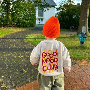 Puede incluir: Un gorro de punto naranja brillante combinado con una sudadera de manga larga color crema que presenta un gráfico rectangular en la espalda con rayas onduladas rosas, amarillas y naranjas y el texto GOOD MOOD CLUB en rojo, junto a pantalones verdes a cuadros.