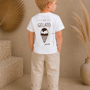 Può includere: Un bambino indossa una maglietta bianca con una grafica di gelato e la parola "gelato" stampata sul retro. La maglietta misura 12 cm di larghezza e 18 cm di lunghezza. Il bambino indossa anche pantaloni beige e scarpe da ginnastica bianche.