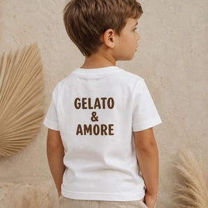 May include: A white t-shirt with the words "GELATO & AMORE" in brown lettering. The shirt is worn by a child, who is facing away from the camera. The child is wearing beige pants.