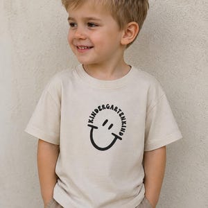 Könnte beinhalten: Ein beiges kurzärmeliges T-Shirt mit einem schwarzen Grafikdruck auf der Brust. Das Design besteht aus einem kreisförmigen Schriftzug KINDERGARTENKIND, der ein einfaches, handgezeichnetes Smiley-Gesicht mit zwei Augen und einem Mund umschließt.