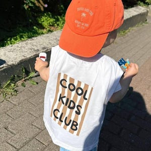 Puede incluir: Una gorra de béisbol naranja con el texto "SAMBA INTO THE NIGHT OF RIO EST. 1976" y un icono de palmera, junto a una camiseta blanca sin mangas con un rectángulo de rayas marrones y blancas con el texto negro "COOL KIDS CLUB" en la espalda.