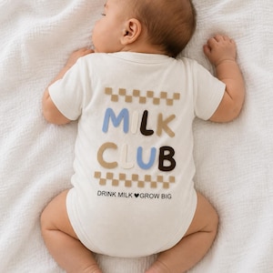 Könnte beinhalten: Ein cremefarbener Kurzarm-Body mit dem Schriftzug MILK CLUB in blauen, braunen und beigen Buchstaben, eingerahmt von zwei Reihen Schachbrettmuster. Unter dem Text ist der Satz DRINK MILK HEART GROW BIG in Schwarz aufgedruckt.