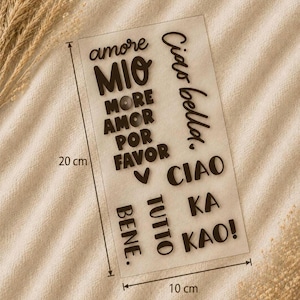 May include: A rectangular clear sheet featuring black text decals including the phrases amore MIO, MORE AMOR POR FAVOR, Ciao bella, CIAO, TUTTO BENE, and KA KAO, with a visible measurement of 20 cm by 10 cm indicated by black lines and arrows.
