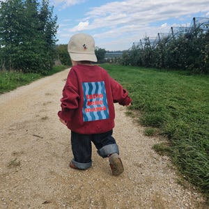 May include: A beige baseball cap with a small Mickey Mouse graphic, a maroon long-sleeve sweatshirt featuring a blue wavy rectangular print with the red text "tutto bene mama", and dark pinstriped trousers with cuffed hems and brown shoes.