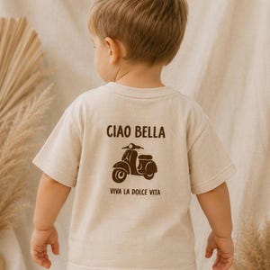 Könnte beinhalten: Beiges T-Shirt für Kinder mit braunem Motorroller-Motiv und den Aufschriften "CIAO BELLA" und "VIVA LA DOLCE VITA". Das Shirt wird von einem kleinen Kind getragen, von hinten gesehen.