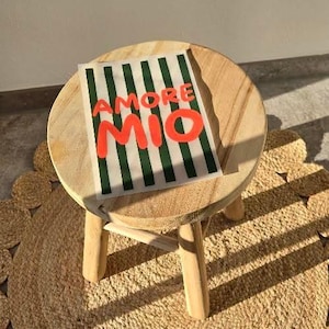 Könnte beinhalten: Ein kleiner, runder, heller Holzhocker mit drei Beinen. Auf der Sitzfläche liegt ein rechteckiger Druck mit vertikalen dunkelgrünen Streifen und dem Text AMORE MIO in fetten, leuchtend orangen Großbuchstaben in der Mitte.