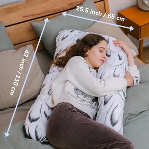 May include: A white and black patterned body pillow, 43 inches (110 cm) long and 25.5 inches (65 cm) wide, is shown on a bed with sage green bedding. The pillow is being embraced by a person wearing a white sweater and brown pants.