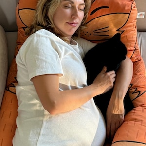 May include: A woman is sleeping on a large orange and brown fox-shaped pillow with a black cat in her arms. The pillow has a black and white design and the woman is wearing a white t-shirt and black shorts.