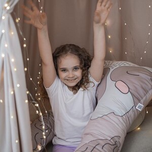 Pode incluir: Uma menina com cabelo encaracolado sorri enquanto está sentada em uma almofada grande de cor marrom claro e rosa com um rosto de desenho animado. A almofada tem a forma de um cilindro longo e fino. A menina está vestindo uma camiseta branca e leggings roxos. O fundo é uma parede marrom claro com luzes brancas.