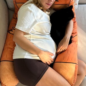 Könnte beinhalten: Eine schwangere Person schläft auf einem großen, orangefarbenen und braunen Kissen in Form eines Fuchses. Die Person trägt ein weißes T-Shirt und schwarze Shorts. Das Kissen ist weich und flauschig.