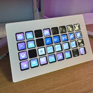 以下が含まれることがあります： 複数の光る正方形のボタンが付いた、銀色のコントロールパネル。各ボタンには、アプリやソーシャルメディアのロゴなど、ユニークなアイコンが表示されています。パネルは木製の表面に置かれています。