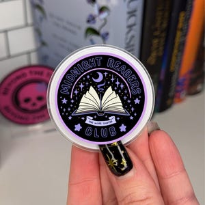 Puede incluir: Una pegatina redonda de color morado y negro con el texto "Midnight Readers Club" y una luna creciente y un libro abierto con el texto "One More Chapter" sobre un fondo negro.