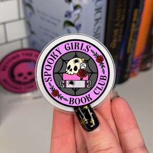 Op de afbeelding: Een ronde sticker in roze en zwart met de tekst "Spooky Girls Book Club" en een schedel met een spin op een boek met een kaars en een roos.