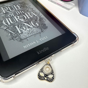 Può includere: Un e-reader Kindle con una custodia trasparente mostra il libro "Rule of the Aurora King" di Nisha J. Tuli. Un ciondolo a forma di gatto nero e oro è appeso al dispositivo. Il ciondolo presenta un disegno di sole e occhi di gatto.