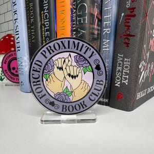May include: A purple and white round book club sign with the text "Forced Proximity Book Club" and a graphic of three hands holding each other with black nail polish. The sign is on a clear acrylic stand.
