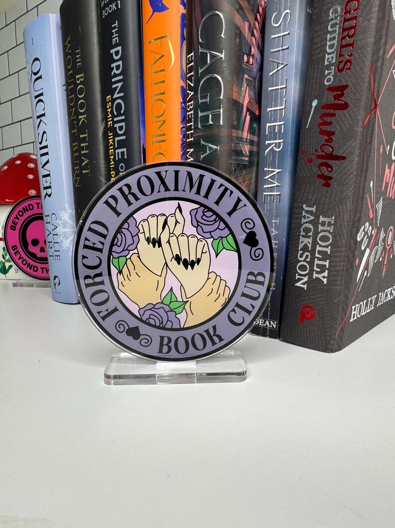 May include: A purple and white round book club sign with the text "Forced Proximity Book Club" and a graphic of three hands holding each other with black nail polish. The sign is on a clear acrylic stand.