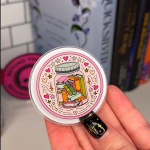 Könnte beinhalten: Ein runder, klarer Aufkleber mit einem Glas voller bunter Bücher. Das Glas ist rosa umrandet, mit rosa Herzen und Sternen am Rand. Der Aufkleber wird vor einem verschwommenen Hintergrund von Büchern gehalten.