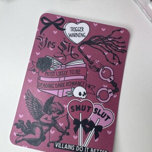 Op de afbeelding: Een rechthoekige, bordeauxrode kaart met verschillende illustraties en tekst. De kaart toont een cupido, handboeien, een zweep en de woorden "Yes Sir", "Trigger Warning" en "Smut Slut". De zin "Villains Do It Better" is ook aanwezig.