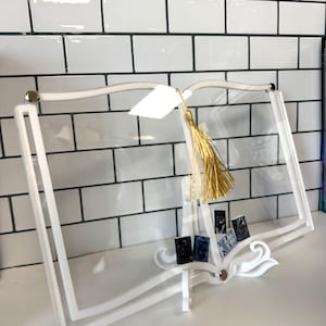May include: A white acrylic book-shaped display stand with a clear center, featuring a gold tassel and small decorative books. The stand is designed to resemble an open book, with a decorative base.