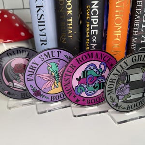 May include: Six round bookish themed stickers with different designs and colors. The stickers have a black outline and the words "Shadow Daddy", "Fairy Smut", "Monster Romance", "Morally Grey", and "Enemies" are printed on them.