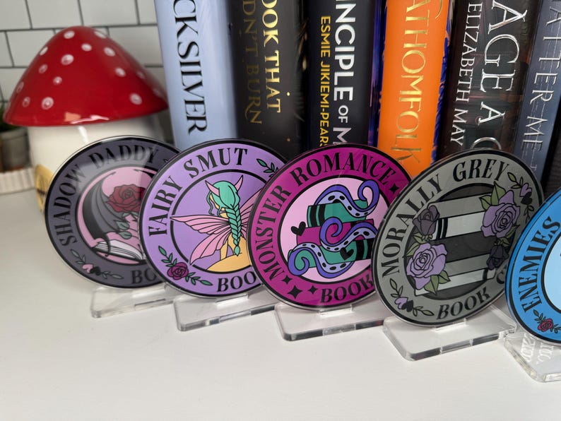 May include: Six round bookish themed stickers with different designs and colors. The stickers have a black outline and the words "Shadow Daddy", "Fairy Smut", "Monster Romance", "Morally Grey", and "Enemies" are printed on them.