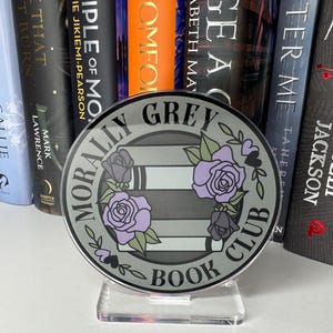 May include: A round, gray bookend with the text "Morally Grey Book Club" in black lettering. The bookend features a stylized design of stacked books with purple roses and black spade symbols.