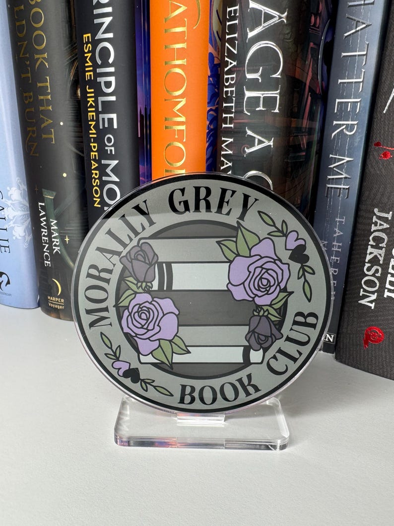 May include: A round, gray bookend with the text "Morally Grey Book Club" in black lettering. The bookend features a stylized design of stacked books with purple roses and black spade symbols.