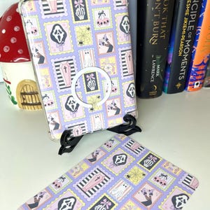 Puede incluir: Una funda de tableta lavanda y una funda a juego presentan un patrón repetido de sellos espeluznantes con fantasmas, arañas y manos de esqueleto. La funda tiene un soporte de anillo blanco. El tema es Halloween.