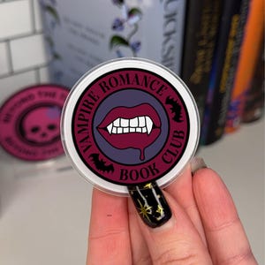 May include: A round sticker with a purple background and a white circle around it. The circle has the text "Vampire Romance Book Club" in black text. Inside the circle is a red vampire mouth with fangs and dripping blood.