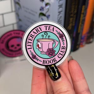 May include: A round sticker with the text "LITERARY TEA BOOK CLUB" encircling a pink teacup with a tea bag. The teacup is set against a teal background, with rose illustrations. The sticker is held in front of a blurred background of books.