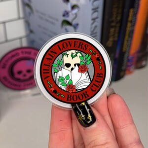 May include: A round sticker with a red border and the text "Villain Lovers Book Club" in white. A skull with vines growing from it is in the center of the sticker, with a book and red roses behind it.
