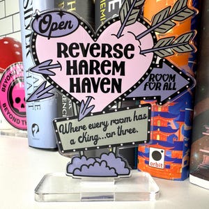 Könnte beinhalten: Ein klarer Acrylschild mit einem rosa Herz, das von Pfeilen durchbohrt wird. Auf dem Schild steht "REVERSE HAREM HAVEN" mit "Open" und "ROOM FOR ALL" auf separaten Schildern. Darunter steht "Where every room has a king...or three."