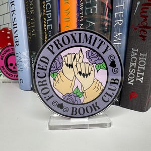 May include: A round acrylic book club sign with the text "Forced Proximity Book Club" and an illustration of two hands holding each other with flowers and hearts. The sign is purple and black.