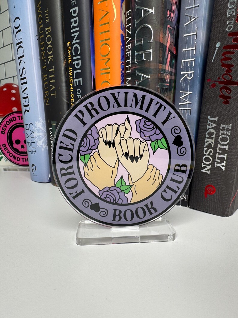 May include: A round acrylic book club sign with the text "Forced Proximity Book Club" and an illustration of two hands holding each other with flowers and hearts. The sign is purple and black.