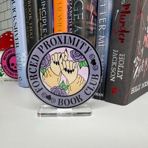May include: A purple and white round sign with the text "Forced Proximity Book Club" and an illustration of two hands holding each other with a rose behind them.