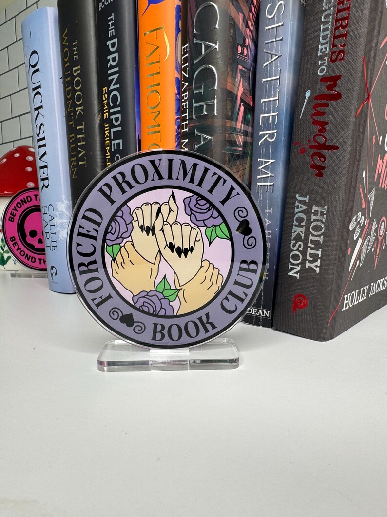 May include: A purple and white round sign with the text "Forced Proximity Book Club" and an illustration of two hands holding each other with a rose behind them.