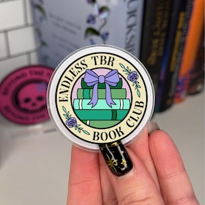 Könnte beinhalten: Ein klarer, runder Aufkleber mit dem Text "ENDLESS TBR BOOK CLUB", der einen Stapel grüner Bücher umrandet, die mit einer lila Schleife zusammengebunden sind. Das Design umfasst kleine lila Rosen und Blätter.