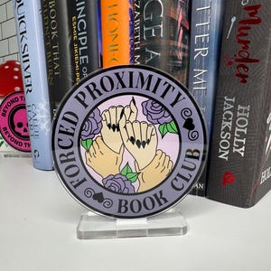 May include: A purple and black round acrylic stand with the text "Forced Proximity Book Club" and a graphic of hands holding each other with roses around them.
