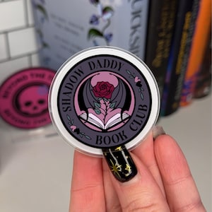 Puede incluir: Una pegatina redonda con fondo morado y borde blanco. La pegatina presenta un murciélago negro con las alas extendidas, una rosa rosa y un libro. El texto "Shadow Daddy Book Club" está escrito en un círculo negro alrededor de la imagen.