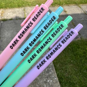 Può includere: Un gruppo di penne dai colori pastello, rosa, blu, verde e viola. Ogni penna presenta la scritta "DARK ROMANCE READER" in nero. Le penne sono disposte a ventaglio, mettendo in risalto i colori e il testo.