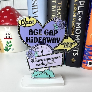 May include: A decorative sign featuring the text "AGE GAP HIDEAWAY" in a heart shape, with the words "Where experience meets youth." The sign is lavender, black, and white, with floral accents and a clock design. The sign is on a white base.