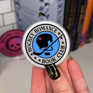May include: A white round sticker with a black border and the text "Hockey Romance Book Club" in a circle around a blue circle with a black silhouette of a hockey player wearing a helmet with a red star on it. The player is holding a hockey stick.
