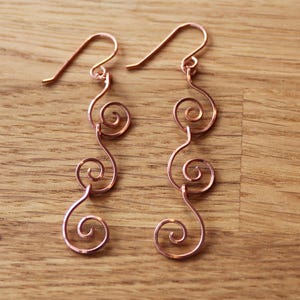 May include: A pair of copper-coloured wire earrings with a swirling design. Each earring has a hook and a series of connected spirals, creating a flowing, elegant shape. The earrings are displayed on a wooden surface.