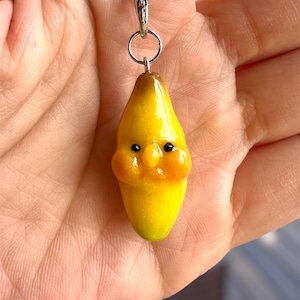May include: A yellow banana keychain with a cute cartoon face. The banana has black eyes, a small red mouth, and rosy cheeks. The keychain is attached to a silver metal ring.