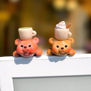 May include: Two small bear figurines, one orange and one tan, each featuring a cup balanced on its head. The orange bear supports a plain cream-colored cup, while the tan bear supports a cream-colored cup topped with white whipped cream.