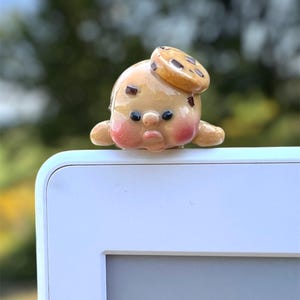 May include: A small, glossy, tan ceramic figurine shaped like a round character with rosy pink cheeks, dark eyes, and a small mouth. The character has a flat, circular cookie-like piece resting on its head and is perched on a white edge.