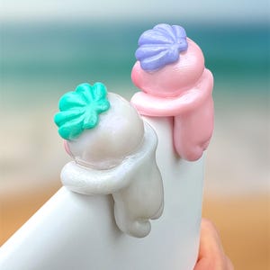 May include: Two colorful phone grips in the shape of hugging figures. One is white with a teal accent, the other is pink with a purple accent. The grips are attached to a white phone, with a blurred ocean background.