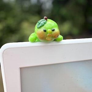 May include: A whimsical, green, pear-shaped figurine with a small leaf and a red dot on top. The figurine has rosy cheeks and is perched on a white electronic device. The background is a blurred view of green foliage.