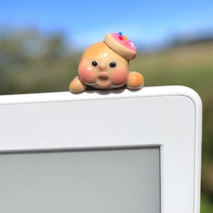 May include: A whimsical, handmade figurine of a smiling, peach-colored face with rosy cheeks and a surprised expression. It wears a small donut hat with pink frosting and colorful sprinkles. The figurine is perched on a white surface against a blurred outdoor background.